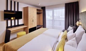 A modern hotel room with a large bed, a television, and a wooden wardrobe. The windows are draped with curtains, and the room has a bright, inviting atmosphere.
