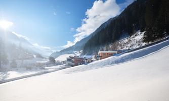 A snowy landscape with houses in the valley. The sky is clear and blue, surrounded by mountains and forests.