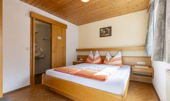 A cozy bedroom with a large bed and wooden ceiling. The walls are bright, and there are natural light sources as well as a small seating area.