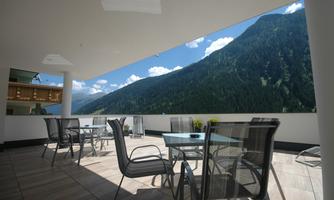 A modern terrace with tables and chairs, surrounded by a beautiful mountain landscape. The sky is clear and blue, creating an inviting atmosphere.