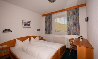 A simple and cozy bedroom with two beds and colorful curtains. A small table and a plant complement the decor.