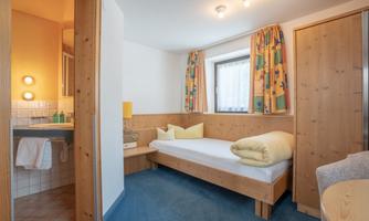 A simple room with wooden ceilings and colorful curtains. There is a single bed, a table, and a sink in the corner.
