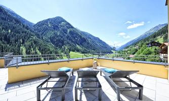 A sunny terrace with loungers and a view of the majestic mountains. The landscape is green and inviting, perfect for relaxing.