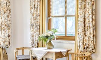 A cozy dining area with a small table and two chairs. The windows are adorned with decorative curtains, and a flower vase sits on the table.