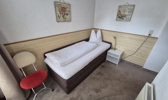 A simple bedroom with a single bed, a chair, and a nightstand. The walls are light and adorned with decorative pictures.