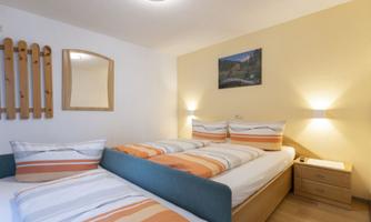 A cozy bedroom with two single beds, decorated with colorful bedding. The walls are painted in a light yellow, and there is pretty lighting.