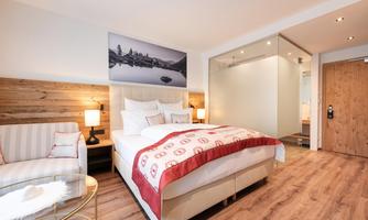 A modern hotel room with a double bed and a cozy seating area. The walls are light, and a large window provides plenty of daylight.