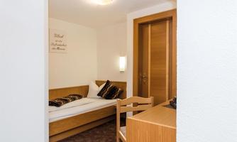A simple room with a bed, a table, and a chair. The walls are light and there are wooden details.
