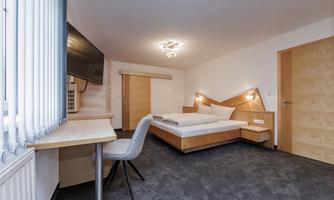A modern hotel room with a double bed and a desk. The room is bright and welcoming, with a large window and simple design.