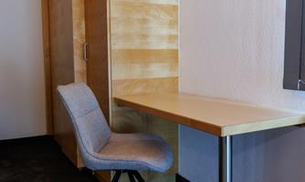 A simple workspace with a bright table and a comfortable chair. In the background, a wooden cabinet can be seen.