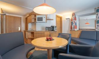 A cozy living area with a small kitchen and a round table. The seating is blue, and there are some apples on the table.