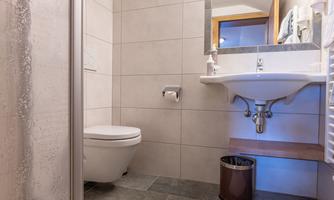 A modern bathroom with a shower, a sink, and a toilet. The floor is tiled, and there is a small doormat.