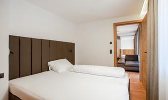 A modern bedroom with a large bed and neutral colors. In the background, a living room with a couch is visible.