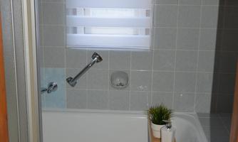 A modern bathroom with a bathtub and a shower cabin. There is a window blind and a small plant on a wooden shelf.