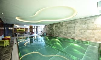 An elegant pool area with greenish shimmering water and modern design. The walls are made of light tiles, and the lighting creates a relaxed atmosphere.
