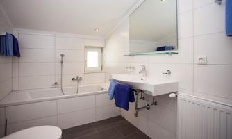 A modern bathroom with white tiles and a bathtub. The amenities include a sink with a mirror and towels in blue color.