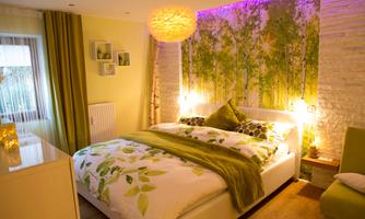 A cozy bedroom with a large bed and green bedding. The wall is decorated with a beautiful forest motif, and soft light creates a relaxing atmosphere.
