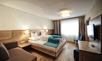 A modern hotel room with a double bed and a cozy seating area. Plenty of daylight through the large window and warm wood paneling create an inviting atmosphere.