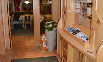 An inviting reception area with a wooden shelf for brochures and a large plant container. In the background, tables and chairs can be seen waiting for guests.