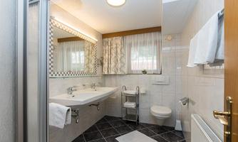 A modern bathroom with tiled floor and large windows. There are two sinks, a toilet area, and towels on the wall.