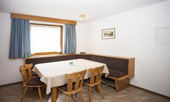 A simple dining area with a table and chairs. The room is bright and has a window with curtains.