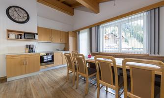 A modern kitchen with a large dining table and wooden chairs. An idyllic mountain landscape is visible through the windows.