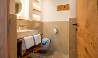 A modern bathroom with a sink, towels, and wooden shelves. The walls are in neutral tones and there is a round light above the sink.
