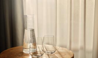 A glass of water and two glasses are on a wooden table. Soft light falls through the curtains.
