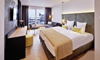 A modern hotel room with two beds and a cozy seating area. Large windows offer a view of the surroundings.