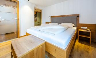 A modern bedroom with a large bed and wooden flooring. The walls are light and there is a nightstand next to the bed.