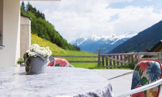 A beautiful table on a terrace with colorful chairs and a view of the mountains. The sky is clear and there is a green meadow in the background.