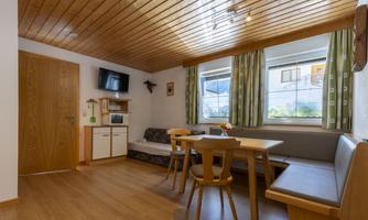 A cozy holiday apartment with wooden ceilings and large windows. The room is simply furnished, with a dining table, chairs, and a television.
