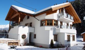 A modern, white house with wood paneling, surrounded by snow. In the background, snow-covered mountains and forests can be seen.