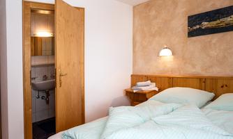 A cozy bedroom with a double bed and soft colors. In the background, there is a bathroom with an open door.