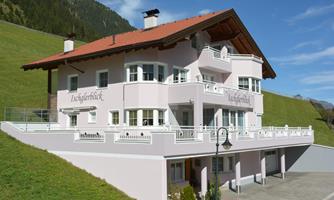 A modern, pink house with a balcony and many windows stands in a green, mountainous setting. The sky is clear and blue.