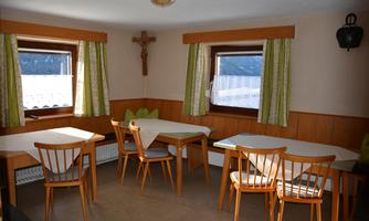 A simply furnished room with wooden tables and chairs. The windows have green curtains and a cross hangs on the wall.