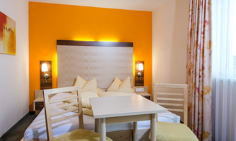 A cozy room with an orange accent and a double bed. The room features a small table and chairs, as well as soft lighting.