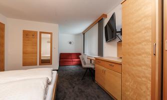 A modern hotel room with a double bed and a gray carpet. There is a television, a desk, and a red sofa.