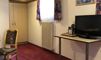 A simple room with carpet, a table, and a television. There is a wardrobe and a window with curtains.