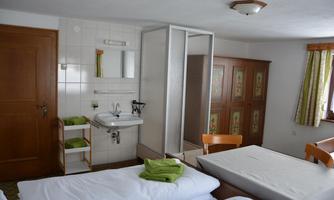 A simple room with two beds, a sink, and a shower area. The furnishings include a wooden cupboard and green towels on the beds.