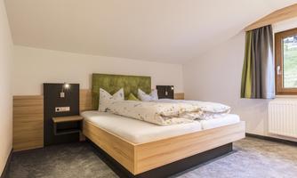 A modern bedroom with a large bed and two pillows. The walls are light, and the windows have curtains in green and gray tones.