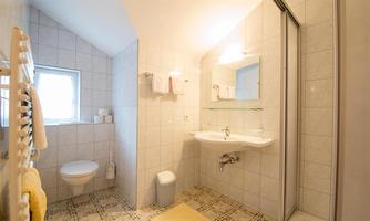A bright bathroom with white tiles and natural light. It features a shower, a toilet, and a sink unit.