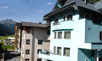 A modern apartment building in a mountain landscape. The surrounding nature features green meadows and majestic mountains in the background.