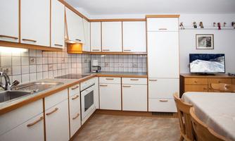 A modern kitchen with white cabinets and a light wood finish. On the table, there is a tablecloth, and a television is visible in the background.