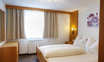 A bright bedroom with a double bed and elegant wooden furniture. Light curtains hang in front of the window, and a picture with floral motifs is hanging on the wall.