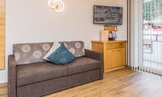 A cozy living room with a sofa and a wooden cabinet. Large windows let in plenty of light and provide a view outside.