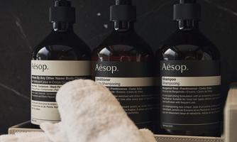 Three bottles of Aesop hair care products are standing on an elegant container. Next to the bottles lie two white towels.
