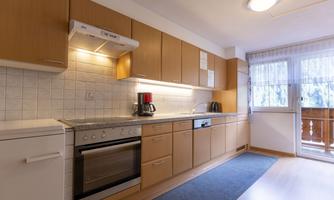 A modern kitchen with wooden cabinets and a refrigerator. Lots of light comes through the window that leads to a balcony.