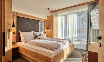 A cozy bedroom with a large bed and wooden furniture. The windows let in plenty of daylight and offer a view outside.