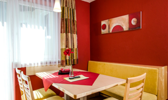 A cozy dining area with a yellow bench and a table, decorated with a red tablecloth. The walls are painted red, and a stylish painting adorns the wall.